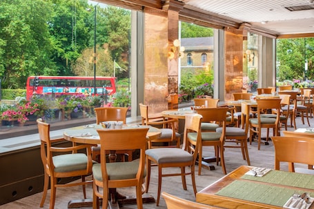 A restaurant with wooden tables and chairs, large windows with a view of a red bus and greenery outside.