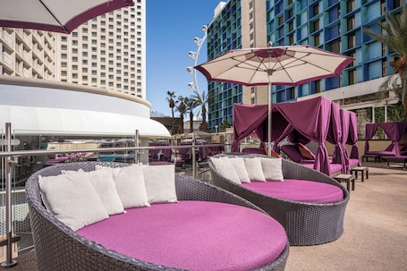 A rooftop lounge area with wicker chairs and purple cushions, overlooking a pool and a high-rise building.