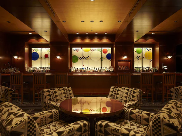 A bar area with a round table surrounded by patterned chairs, wooden paneling on the walls, and a bar counter with bottles.