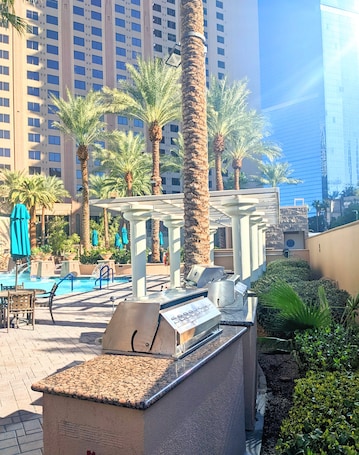 A rooftop pool area with palm trees, a barbecue grill, and a view of high-rise buildings.