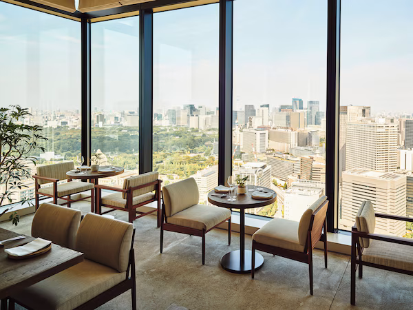 A high-rise hotel room with large windows offering a city view, a round dining table with chairs, and a potted plant.