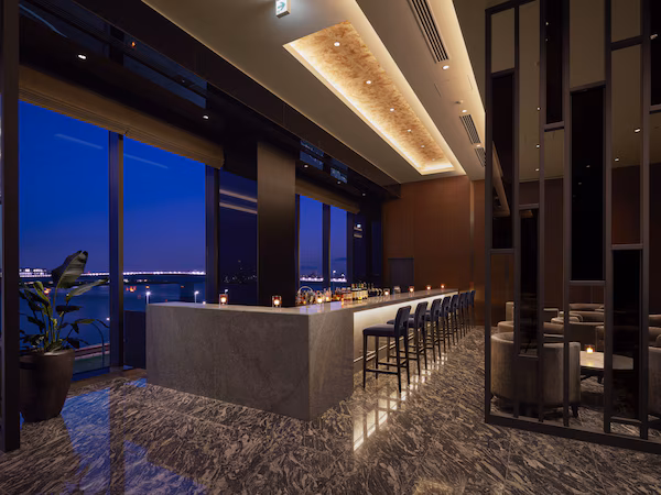 A modern bar area with a marble floor, large windows, and a view of the cityscape at dusk.