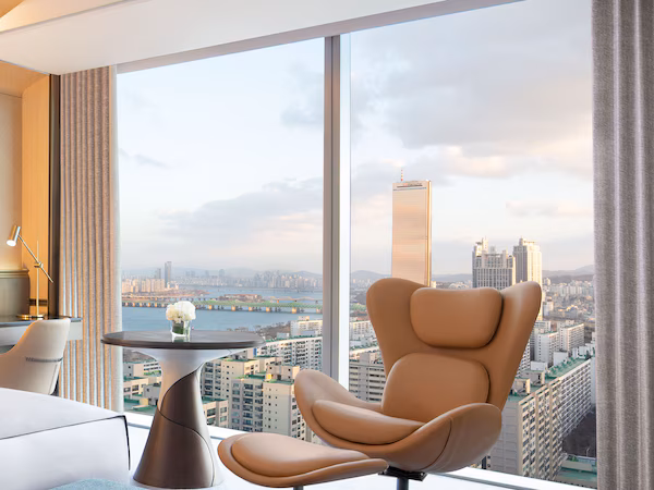 A modern hotel room with a large window offering a city view, a comfortable chair, a small table with a vase, and a lamp.