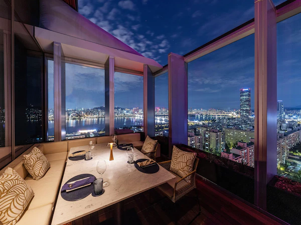 A dining area with a table set for two, large windows offering a city view, and a modern interior design.